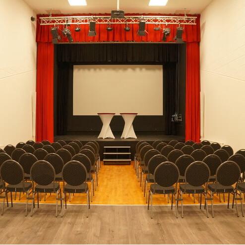 Leerer Veranstaltungsraum mit Reihen bequemer Stühle und Bühne mit zwei Stehtischen und einer Leinwand.