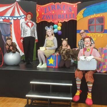 Sechs Personen in Kostümen stehen auf einer Bühne vor einem Zirkushintergrund mit bunten Dekorationen.
