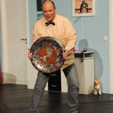 Ein Mann in einem beigefarbenen Hemd hält mit beiden Händen ein großes, rundes Schild auf einer Bühne.