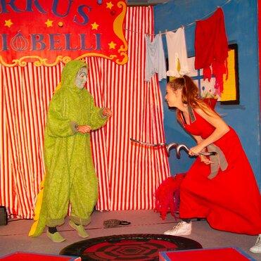 Zwei Schauspieler auf einer Zirkusbühne: einer in grünem Kostüm, der andere in rotem Kleid mit einem Haken.