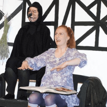 Zwei Schauspieler auf einer Bühne: Eine Frau mit Buch und eine maskierte Figur im Hintergrund vor einer verzweigten Wand.