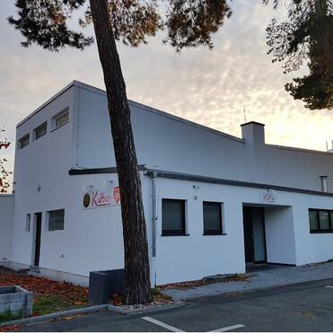 Modernes, weißes Gebäude mit großen Fenstern und einem Eingangsschild, umgeben von Bäumen und herbstlichem Laub.