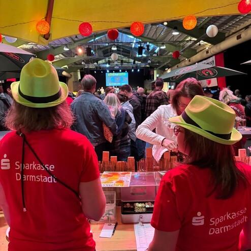 Zwei Personen in roten T-Shirts mit &bdquo;Sparkasse Darmstadt&ldquo; und gelben H&uuml;ten stehen an einem Tisch in einer Menschenmenge.