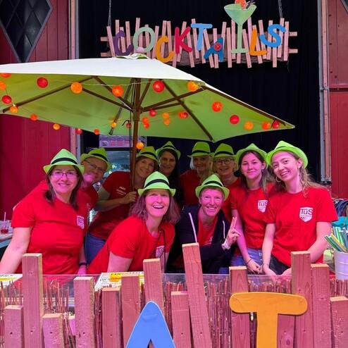 Gruppe von zehn Personen in roten Shirts und gr&uuml;nen H&uuml;ten hinter einem Cocktailstand mit bunten Dekorationen.