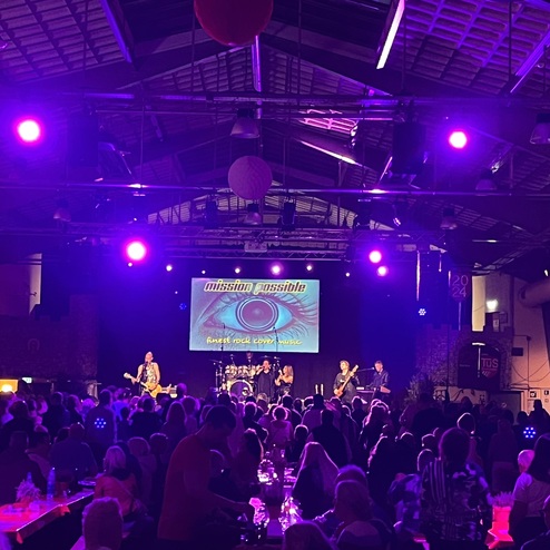 Lebendige Konzertatmosph&auml;re mit Band auf der B&uuml;hne und Publikum in einem beleuchteten Veranstaltungsraum.