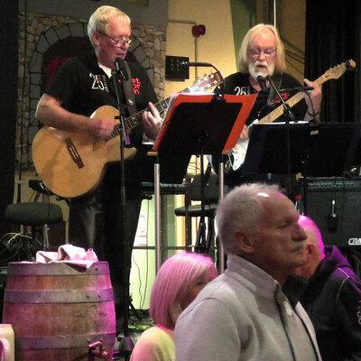 Zwei Musiker mit Gitarre und Bass spielen auf einer B&uuml;hne, w&auml;hrend das Publikum ihnen aufmerksam zuh&ouml;rt.