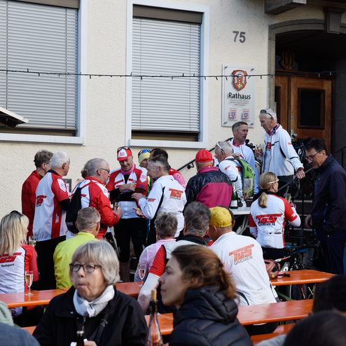 Gruppe von Radfahrern in roten Trikots versammelt sich vor dem Rathaus, w&auml;hrend andere auf B&auml;nken sitzen und reden.