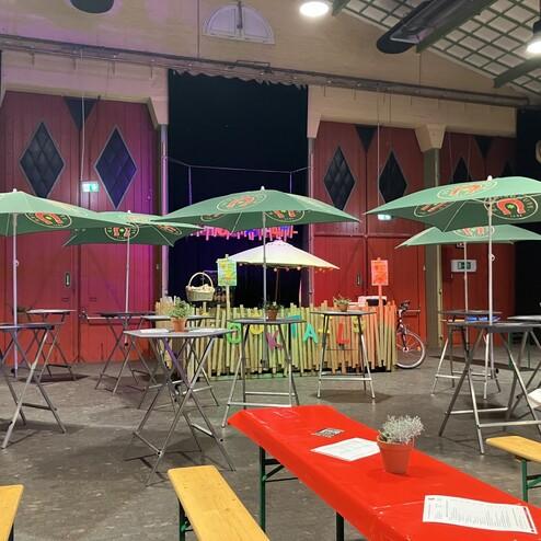 Bunte Sonnenschirme stehen über Stehtischen in einem festlich dekorierten Veranstaltungsraum mit Holzpalisaden.