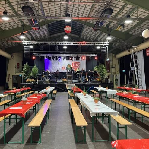 Blick auf eine festlich dekorierte Halle mit Biertischen und einer Band auf der Bühne. Banner mit "Herzlich willkommen".