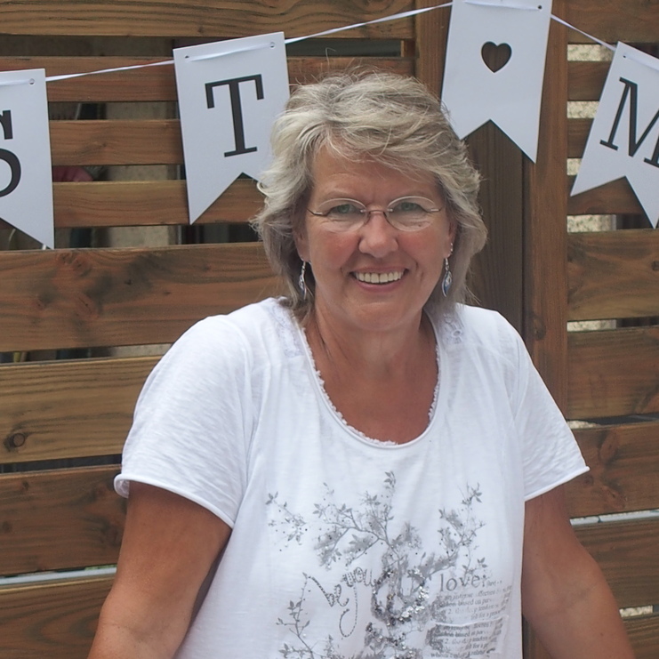 Lächelnde Frau mit kurzen Haaren in weißem T-Shirt, steht an einem Holztisch vor einer Holzwand mit Wimpelgirlande.