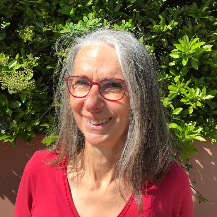 Frau mit langen grauen Haaren und roten Brillenlächeln vor einem üppigen grünen Hintergrund. Sie trägt ein rotes Oberteil.