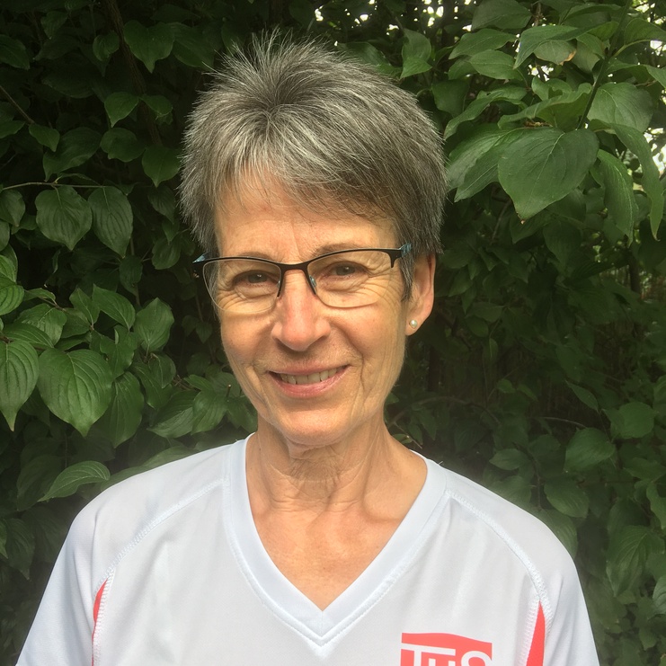 Porträt einer Frau mit kurzem grauem Haar, Brille und T-Shirt mit Aufdruck „TuS Griesheim“ vor grünem Hintergrund.