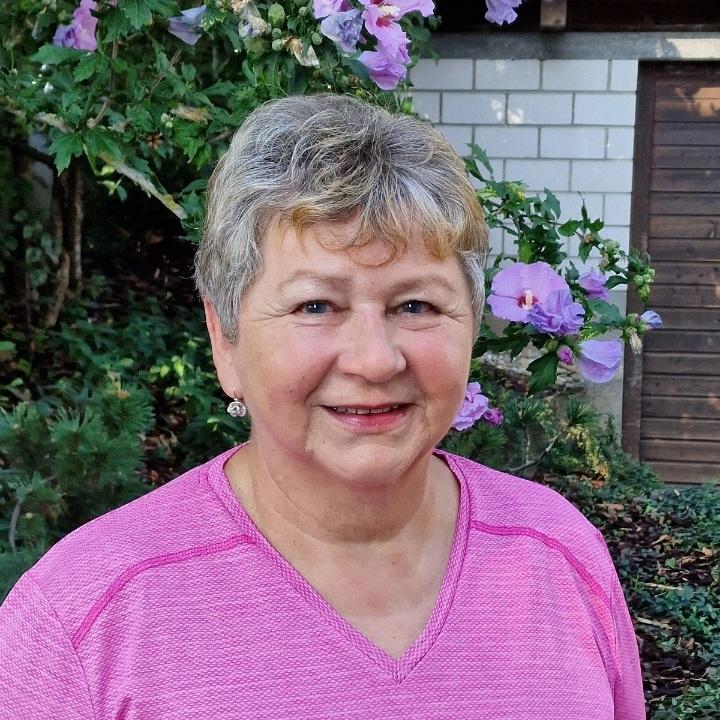 Porträt einer Frau mit kurzen, grauen Haaren in pinkem Shirt vor blühenden Pflanzen und einem Holzschuppen im Hintergrund.