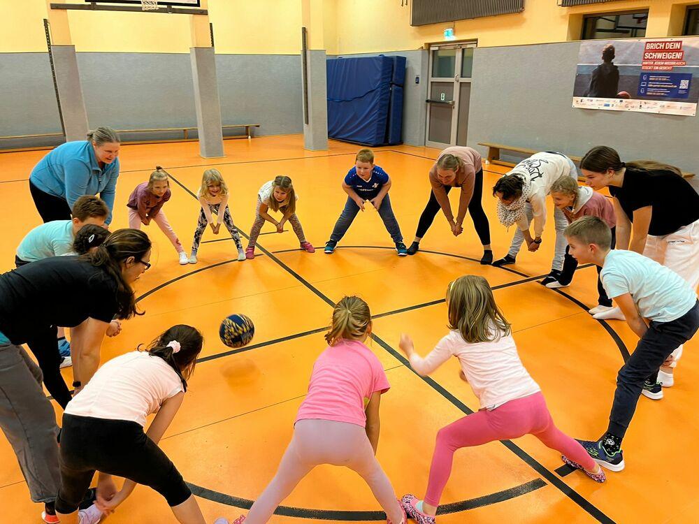 Gruppe von Kindern und Erwachsenen spielt einen Ball auf einem orangefarbenen Sportboden in einer Turnhalle.