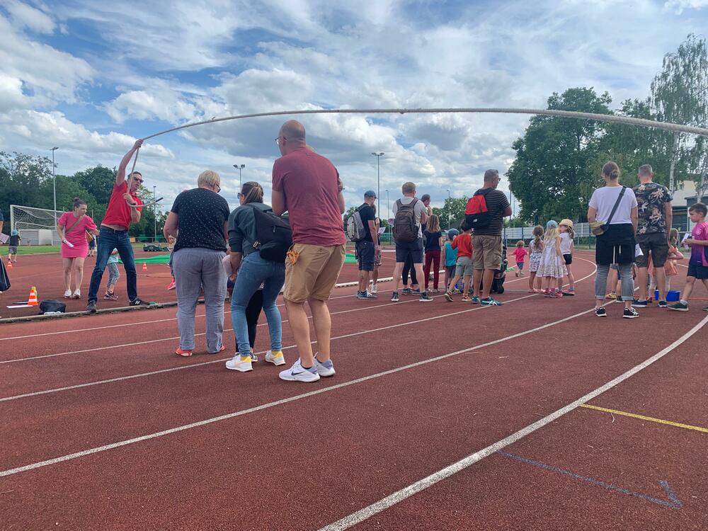 Menschenansammlung auf einem Sportplatz, einige schwingen ein Seil, während Kinder und Erwachsene an einem Spiel teilnehmen.