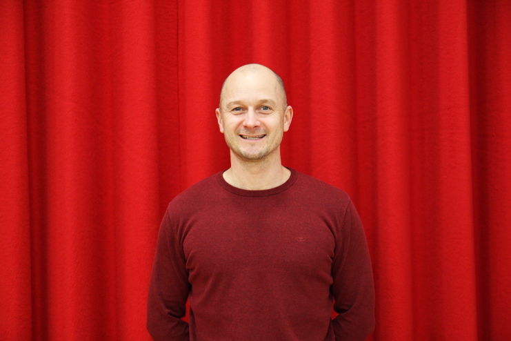 Mann lächelt in einem roten Pullover vor einem Hintergrund aus roten Vorhängen.