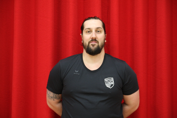 Porträt eines Mannes mit Bart, in schwarzem T-Shirt vor rotem Hintergrund, mit verschränkten Armen.