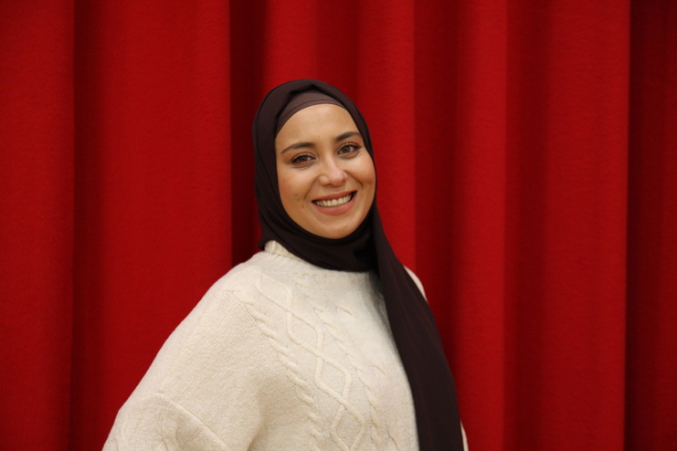 Lächelnde Frau mit braunem Hijab und weißem Pullover vor rotem Hintergrund.