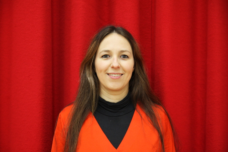 Porträt einer lächelnden Frau mit langen, braunen Haaren, die ein rotes Oberteil und einen schwarzen Rollkragen trägt, vor rotem Hintergrund.