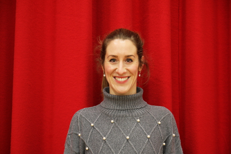 Frau mit lächelndem Gesicht, grauem Rollkragenpullover mit Perlen, vor rotem Hintergrund.