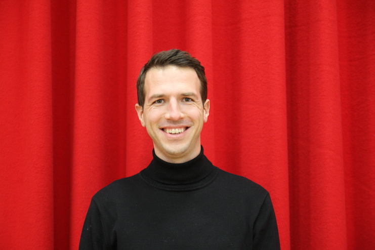 Lächelnder Mann in schwarzem Pullover vor einer roten Stoffwand.