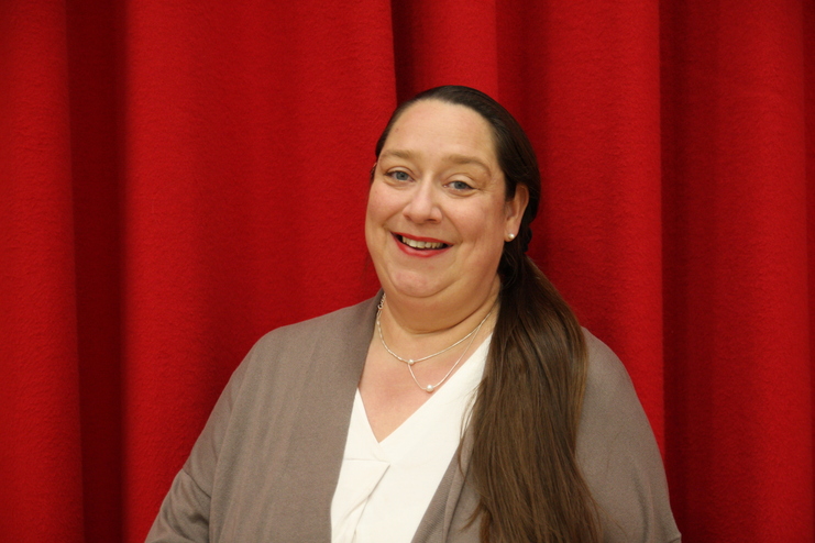 Lächelnde Frau mit langen Haaren steht vor einem roten Stoffhintergrund, trägt einen grauen Cardigan und eine helle Bluse.