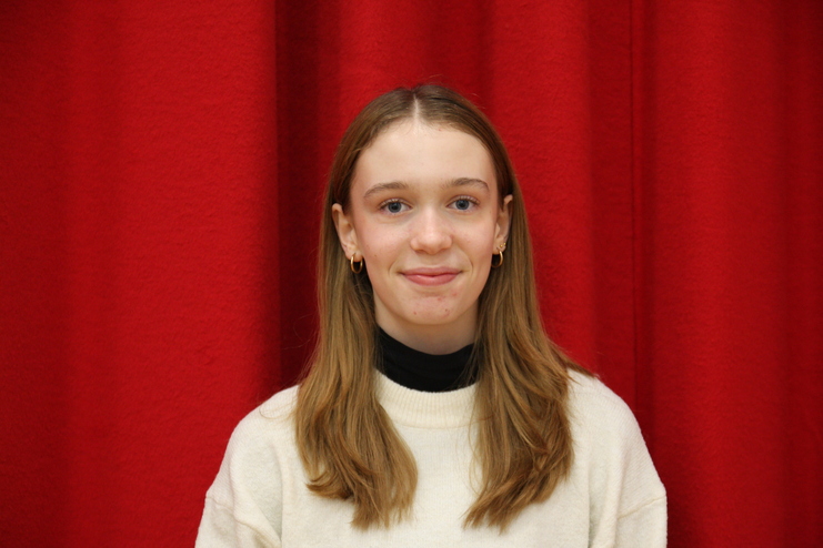 Portr&auml;t einer jungen Frau mit langen, blonden Haaren, die vor einem roten Hintergrund steht und in einem wei&szlig;en Pullover gekleidet ist.