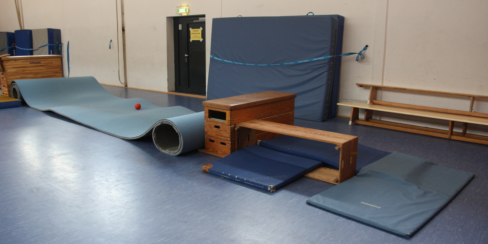 Gymnastikbereich mit einer gerollten Matte, einem Ball und verschiedenen Trainingsger&auml;ten auf dem blauen Boden.