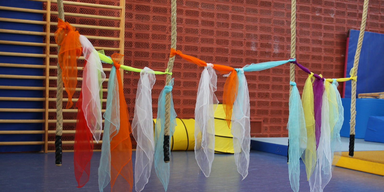 Bunte T&uuml;cher h&auml;ngen an Seilen in einem Turnraum, umgeben von mattenartigen Unterlagen und einer Holzleiste.