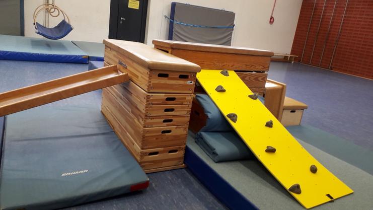 Kletterspielgerät mit Holzplatten, Rutsche und gelber Kletterwand in einem Sportraum mit Matten darunter.