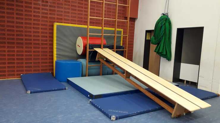 Gymnastikbereich mit einer schrägen Bank, Matten, einem roten und blauen Polster sowie einer Leiter an der Wand.