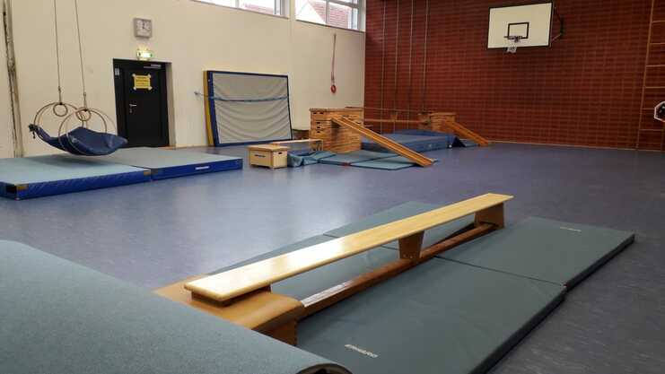 Turnhalle mit Turngeräten: Sprossenwand, Schwebebalken, Matten und Ringe in einem großen Raum mit Holz- und blauen Elementen.