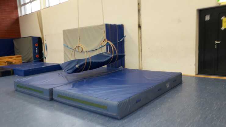 Turnhalle mit blauen Matten, schwingenden Ringen und einem Wandpolster im Hintergrund.