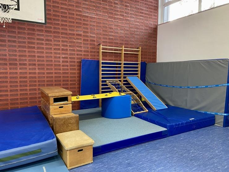 Geräumige Turnhalle mit Klettergerüst, Matten und verschiedenen Gymnastikgeräten an einer Backsteinwand.