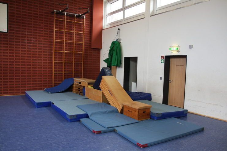 Klettergerüst aus Matten und Holzkisten in einer Turnhalle mit blauen Matten und einer Kletterwand im Hintergrund.