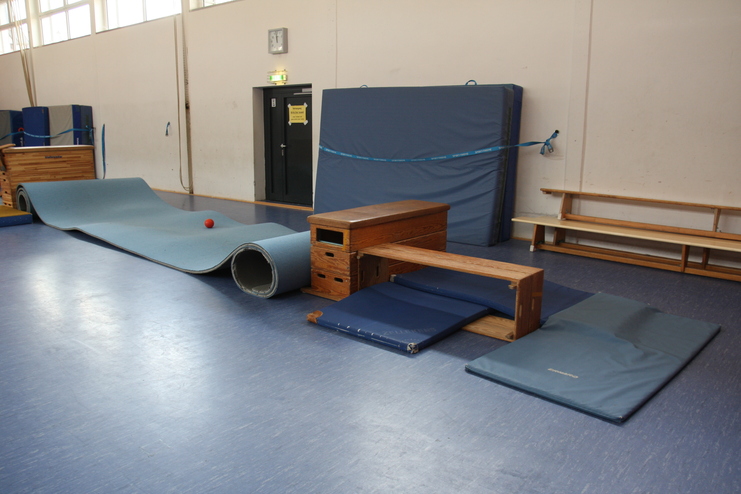 Gymnastikbereich mit einer gerollten Matte, einem Ball und verschiedenen Trainingsgeräten auf dem blauen Boden.