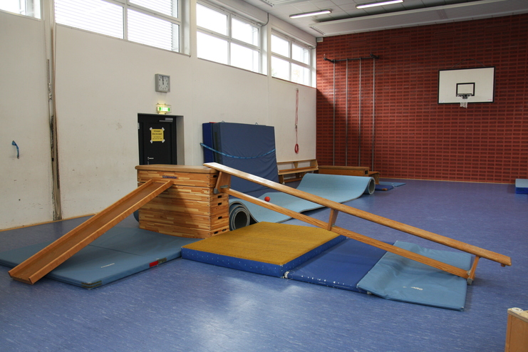 Aufbau aus Turngeräten in einer Sporthalle mit Schrägbrett, Matten und anderen Übungen auf blauem Bodenbelag.