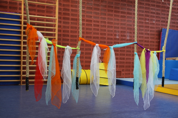 Bunte Tücher hängen an Seilen in einem Turnraum, umgeben von mattenartigen Unterlagen und einer Holzleiste.