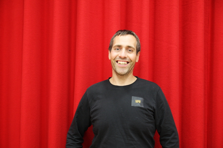 Lächelnder Mann in einem schwarzen Shirt steht vor einem roten Vorhang.
