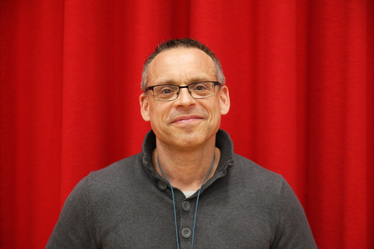 Mann mit Brille und grauem Pullover steht vor einem roten Vorhang und lächelt freundlich in die Kamera.