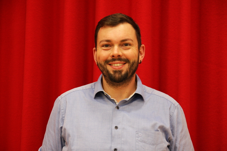 Portrait eines lächelnden Mannes mit Bart, blauer Hemd und rotem Hintergrund.