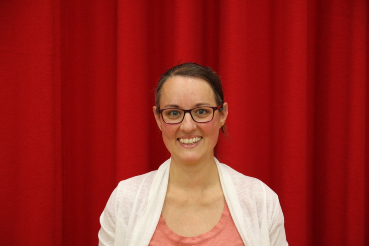 Lächelnde Frau mit Brille vor einem roten Hintergrund, trägt ein helles Oberteil und hat schulterlanges Haar.