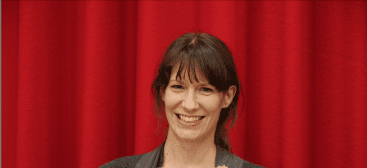 Frau mit lächelndem Gesicht steht vor einem roten Vorhang und hält ein helles Holzobjekt in der Hand.