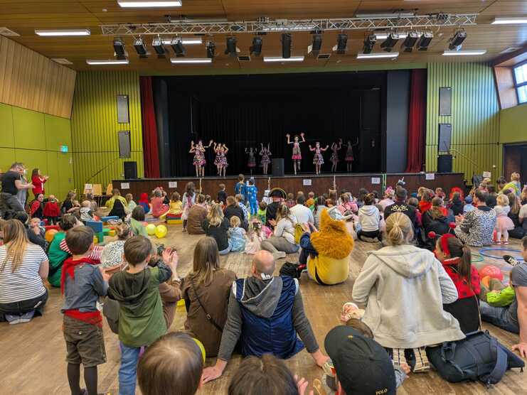 Bunt dekorierte Bühne mit kostümierten Tänzern, während Zuschauer auf dem Boden applaudieren und feiern.
