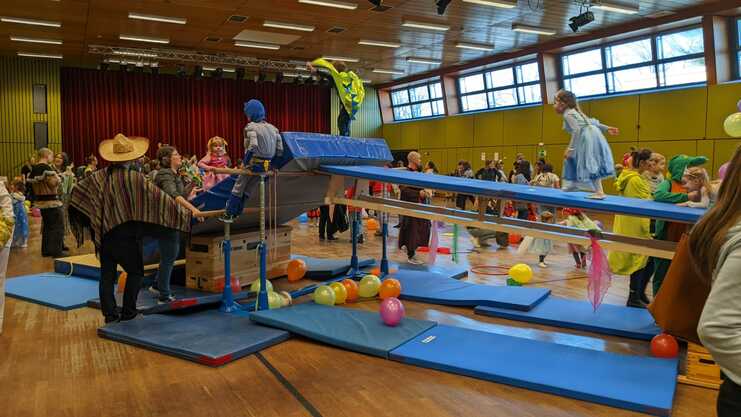 Karnevalsfeier in einer Sporthalle mit spielenden Kindern, Hindernissen und bunten Luftballons auf dem Boden.
