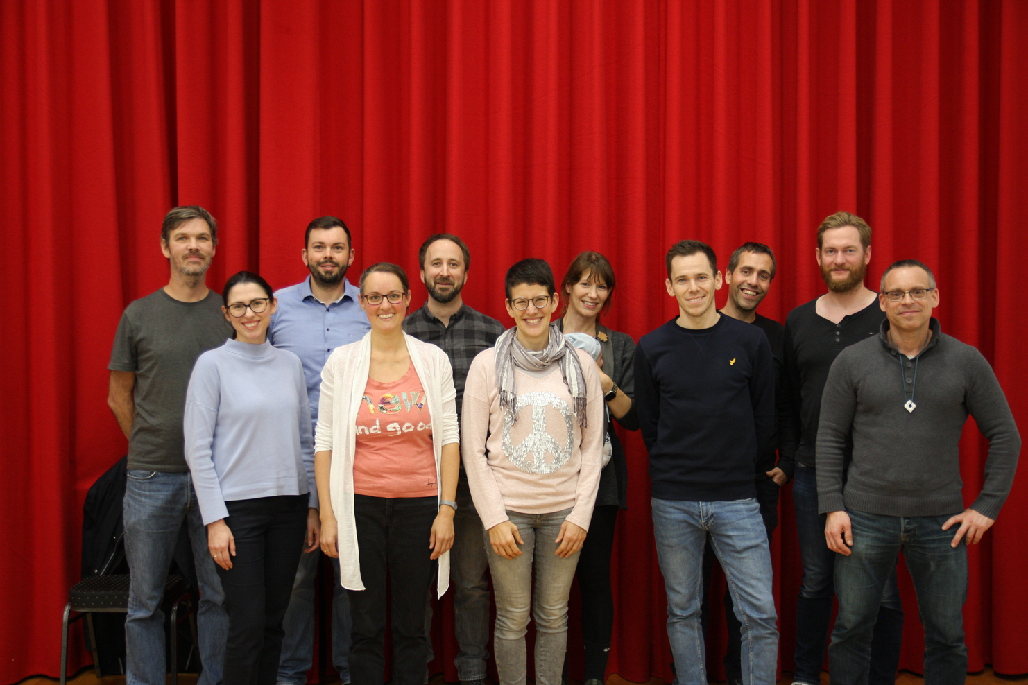 Gruppe von zwölf Personen posiert vor rotem Vorhang, lächelnd und in lockerer Kleidung.