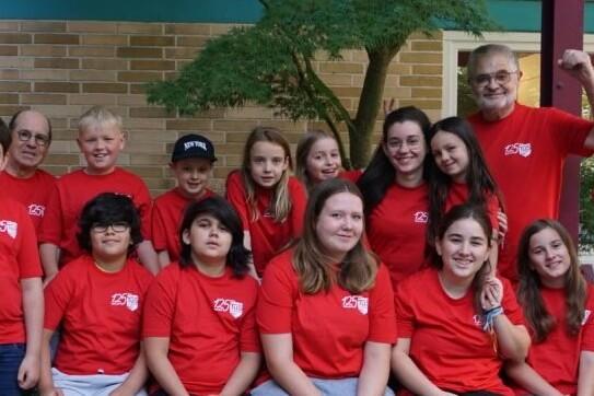 Gruppe von 15 Personen in roten T-Shirts, stehend und sitzend, vor einer Mauer mit Pflanzen im Hintergrund.