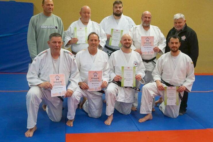 Gruppenfoto von Judo-Prüflingen mit Urkunden auf einer Judomatte, angeführt von einem Trainer im Hintergrund.