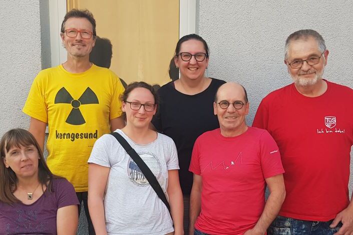 Gruppenfoto von sechs Personen vor einer grauen Wand, alle lächeln und tragen unterschiedliche Kleidung.