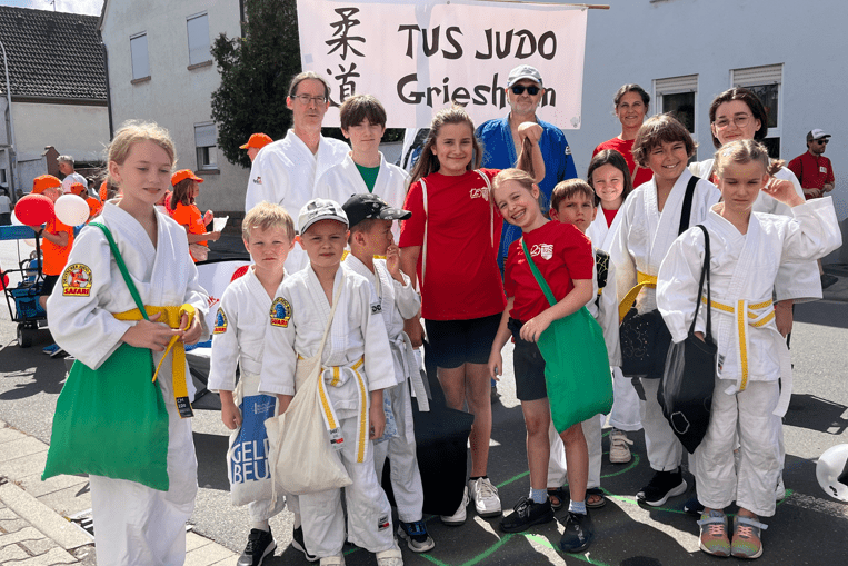 Gruppe von Judokas in weißen Gi, einige mit Gürteln, posieren vor einem Banner mit der Aufschrift „TUS JUDO Griesheim“.